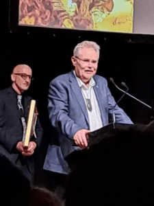 Professor Richie Poulton stands behind a podium with two microphones attached. He has short grey hair and glasses, is wearing a white shirt with a blue jacket and has a greenstone carving around his neck. Another man wearing a black shirt stands in the background holding a golden frame, and the bottom edge of a presentation screen can be seen at the top of the photo.